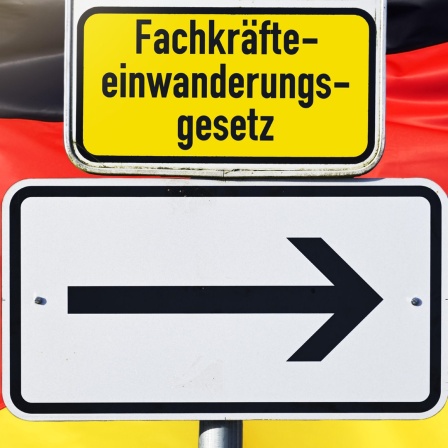 Ein Schild mit einem Pfeil und der Aufschrift Fachkräfteeinwanderungsgesetz vor einer deutschen Flagge