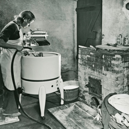 Eine Hausfrau mit Kittel steht im Waschkeller und befüllt eine Miele Waschmaschine in den 50er Jahren.