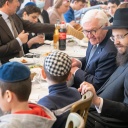 Frank-Walter Steinmeier sitzt mit orthodox gekleideten Juden an einem Tisch beim Essen und spricht mit aus der Ukraine geflüchteten Kindern.