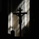 Die Silhouette eines Kreuzes zeichnet sich vor einer spärlich von Sonnenstrahlen angestrahlten Betonwand ab.