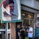 Drei Jugendliche stehen vor einem Lebensmittelgeschäft neben einem Baum mit dem Plakat des iranischen Präsidenten Raisi.
