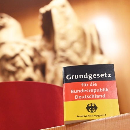 Vor dem Bundesadler im Sitzungssaal des Bundesverfassungsgericht steht eine Miniaturausgabe des Grundgesetz für die Bundesrepublik Deutschland sowie ein Barett eines Bundesverfassungsrichters. 