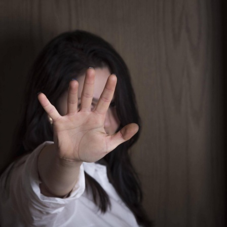 Eine Frau vor einer Holzwand hat ihre Hand zu einer Stopp-Geste ausgestreckt, und verdeckt so ihr Gesicht.