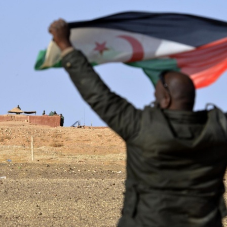Ein Mann aus dem Volk der Sahrauis hält eine Flagge der Widerstandsorganisation Polisario hoch, im Hintergrund sichern marokkanische Soldaten die Grenzmauer zum von Marokko kontrollierten Teil der Westsahara