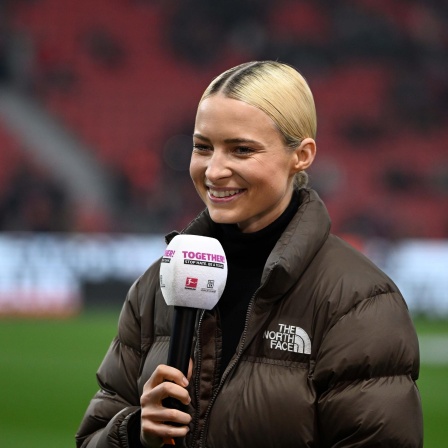 Die Sportmoderatorin Lena Cassel am Mikrofon bei einem Bundesligaspiel im Stadion. Sie trägt einen Rollkragenpullover und eine braune Daunenjacke.