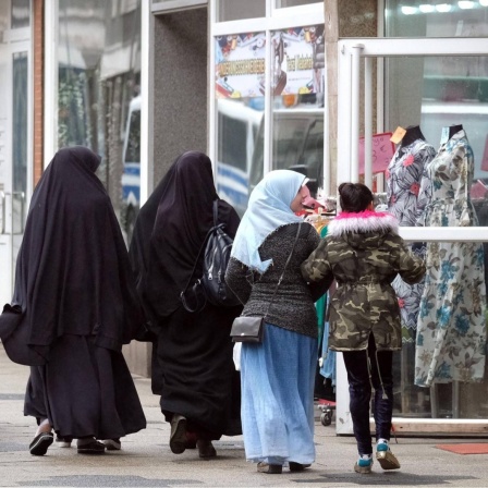 Zwei in schwarz verhüllte ältere Frauen und zwei jüngere Mädchen, davon eins mit mit Kopftuch, passieren ein muslimisches Bekleidungsgeschäft mit Schaufensterpuppen in Duisburg-Marxloh.