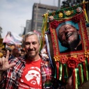 Ein Mann steht in einer Menge auf der Straße und hält ein großes buntumrandetes Foto von Lula da Silva an einem Stock in der linken Hand. Mit der rechten streckt er Zeigefinger und Daumen in die Höhe.