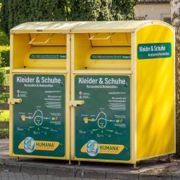 Zwei gelbe Altkleidercontainer mit der Aufschrift "Kleider & Schuhe Accessoires & Heimtextilien" auf einem Parkplatz. 