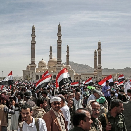 Teilnehmer einer Demonstration halten in Sanaa jemenitische Fahnen in die Höhe, im Hintergrund ist eiine Moschee zu sehen.