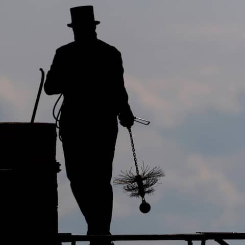 Symbolbild: Ein Schornsteinfeger läuft mit seinem Kaminkehrerwerkzeug auf einem Dach entlang