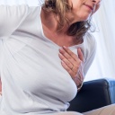 Eine Frau sitzt auf einem Sessel und fasst sich mit schmerzverzerrtem Gesicht an die Brust und den Rücken (gestellte Szene)