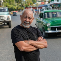 Der kubanische Schriftsteller Leonardo Padura posiert für ein Porträt auf einer Straße in Havanna. Im Hintergrund fährt ein grünes Auto in der für Kuba typischen Anmutung der Fünfzigerjahre.