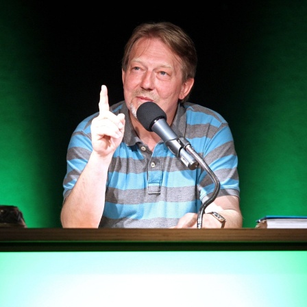Das Bild zeigt Dietmar Wischmeyer, der an einem Tisch sitzt und den rechten Zeigefinger erhoben hat