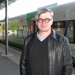 Ein Mann mit einer markanten schwarzen Brille steht vor einem Regionalzug am Bahnhof - es ist der tschechische SchriftstellerJaroslav Rudiš.