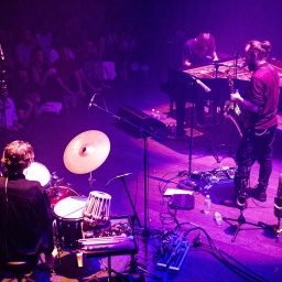 Drei Männer musizieren auf einer Bühne vor Publikum.