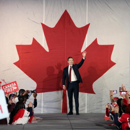 Pierre Poilievre steht bei einer Rally auf der Bühne und winkt. Hinter ihm an der Wand prangt ein riesiges stilisiertes rotes Ahornblatt.