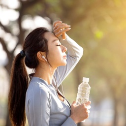 Eine junge, sportliche gekleidete Frau hält sich die Hand an die Stirn, in der anderen Hand hat sie eine Wasserflasche.
