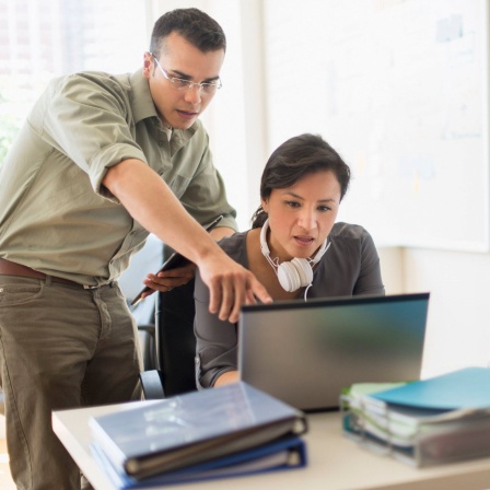 Zwei Menschen arbeiten zusammen an einem Laptop. Die Frau hat Kopfhörer um den Hals hängen (Bild: picture alliance / Bildagentur-online/Tetra Images) 