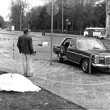 Der Tatort mit den zugedeckten Leichen von Siegfried Buback (vorne links) und seines Fahrers Wolfgang Göbel sowie der Dienstwagen des Generalbundesanwaltes, in dem die beiden erschossen wurde. Der höchste Ankläger der Bundesrepublik ist am 7. April 1977 in Karlsruhe durch Schüsse aus einer Maschinenpistole, die von einem Motorrad aus auf seinen Dienstwagen abgegeben worden waren, getötet worden. Mit ihm starb sein Fahrer Wolfgang Göbel, der lebensgefährlich verletzte Justizhauptwachtmeister Georg Wurster erlag am 13. April seinen Verletzungen.