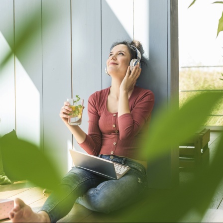 Eine weiße Frau hört mit Kopfhörern Musik, in der Hand ein gesundes Getränk, im Vordergrund unscharfe grüne Blätter