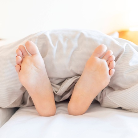 Zwei Füße schauen unter einer Bettdecke hervor: Kinder fallen im Schlaf gelegentlich aus dem Bett, weshalb man sie oft in Gitterbetten legt. Erwachsene dagegen brauchen das nicht. Warum ist das so?