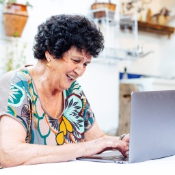 Eine Seniorin sitzt an einem Tisch am Laptop und lacht. 