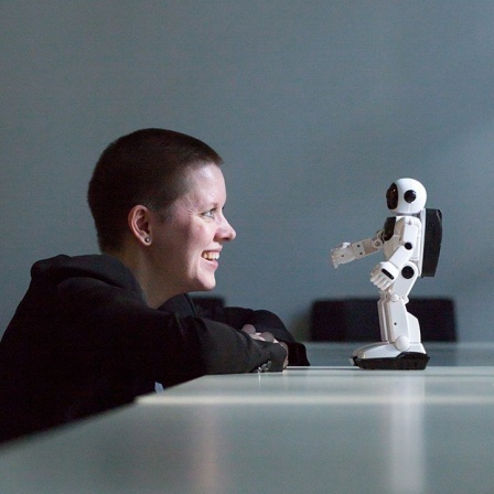 Die Philosophin Janina Loh sitzt am Tisch und blickt auf einen kleinen weissen Roboter ihr gegenüber.