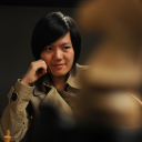 Hou Yifan sitzt vor einem schwarzen Hintergrund und trägt Trenchcoat. Sie blickt herausfordernd auf einen Schach-Springer, der sich unscharf im Vordergrundgrund befindet und perspektivisch das Bild bestimmt