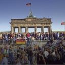 Fall der Berliner Mauer - Menschen sitzen auf der Mauer vor dem Brandenburger Tor, eine große Menschenmenge steht davor