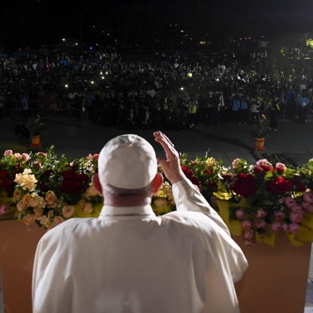 Papst Franziskus steht in Papua-Neuguinea auf einer beleuchteten Bühne vor einer Menschenmenge im Dunkeln, die Lichter in die Höhe hält. 