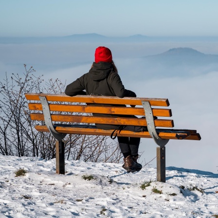 Eine Person sitzt auf einer Bank auf dem verschneiten Staffelberg und blickt in das wolkenbehangene Tal.