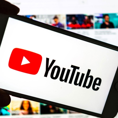 Das Logo des Video-Portals YouTube wird auf dem Display eines Smartphones angezeigt. Im Hintergrund ist auf einem Bildschirm die YouTube Homepage zu sehen
