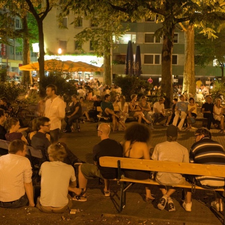 Szenetreff Brüsseler Platz vor der Kirche St. Michael im Sommer. Bild: Menschen stehend, sitzend, erleuchtet