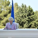 Zwei Personen, eine Frau und ein Mann, sitzen an einem langen weißen Tisch und unterzeichnen Verträge. Im Hintergrund sieht man Bäume, die Flagge der EU und von Aserbaidschan.