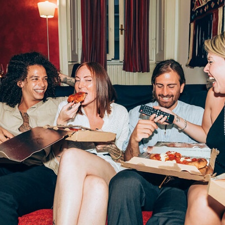 Junge Menschen essen lachend Pizza auf einem Sofa.