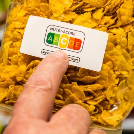 Eine Packung Cornflakes, ein Zeigefinger deutet auf die Farbflächen des Nutri-Score-Aufdrucks. Die mittlere Kategorie "C" (gelb) ist hervorgehoben. 