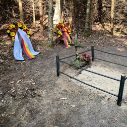 Eine mit Eisengitter umzäunte Grabstelle, im Hintergrund zwei Kränze mit der deutschen und einer hellblauen Flagge.