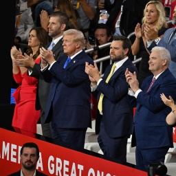 Donald Trump steht mit mehreren Menschen beim Parteitag der Republikaner auf der Tribüne und klatscht. Neben ihm steht J. D. Vance und klatscht ebenfalls. 