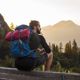 Ein junger Wanderer mit Rucksack macht in den Alpen Pause, sitzt auf einer Bank und blickt auf das Bergpanorama