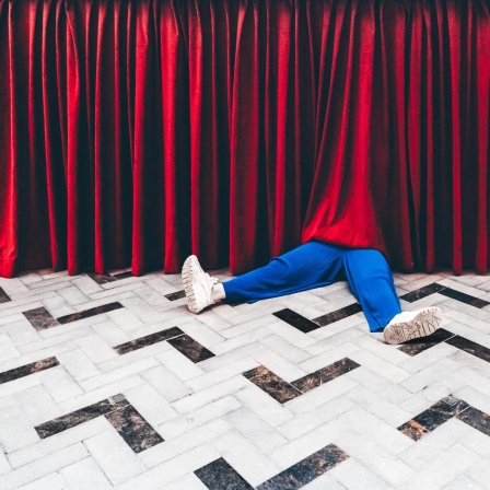 Beine einer Person in blauen Hosen schauen unter einem Theatervorhang heraus.