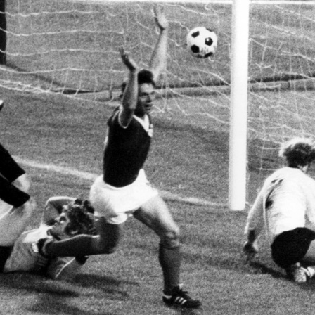 Jürgen Sparwasser reißt jubelnd die Hände hoch, gegenerischer Torwart und Spieler liegen am Boden, der Ball fliegt ins Netz