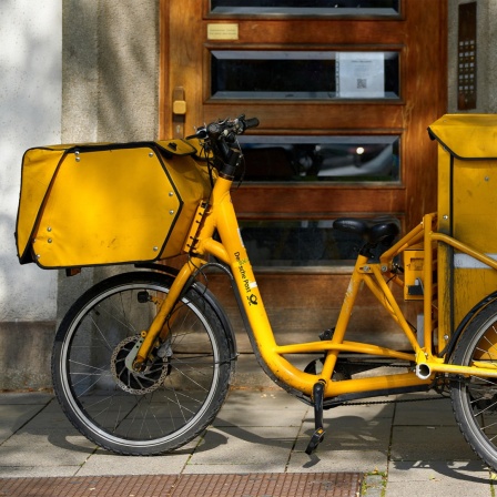 Ein gelbes Postfahrrad der Deutschen Post mit großen Taschen für die Post steht vor einem Gebäude.