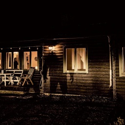 Es ist Nacht, im Holzhaus brennt Licht, auf der Fassade sieht man den großen Schatten eines Mannes.