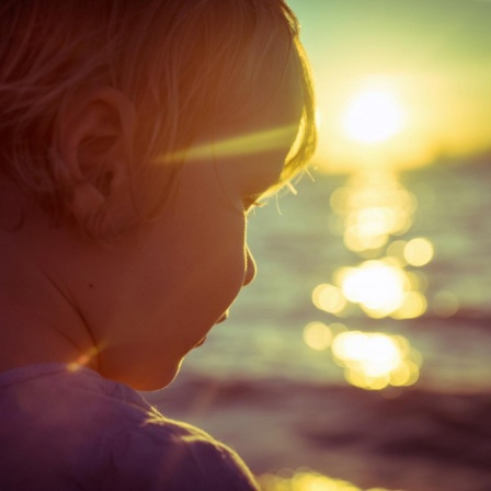 Fotografie eines Kindes am Meer, im Gegenlicht der, am Horizont, untergehenden Sonne.