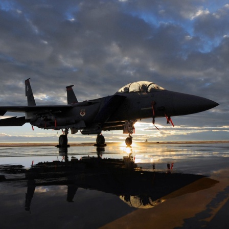 Ein Kampfjet, ein F-15 Strike Eagle fighter, steht zwischen Pfützen auf einem Rollfeld im Sonnenuntergang, im Hintergrund türmen sich dunkle Wolken auf.