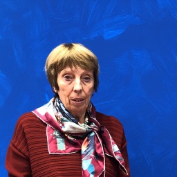 Eine alte Frau in einer weinroten Jacke mit buntem Schal. Sie hat kurze, flachsfarbene, glatte Haare. Hinter ihr ist eine blaue Wand.
