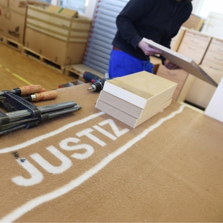 Ein Häftling baut in der Schreinerei der Justizvollzugsanstalt in Ulm (Baden-Württemberg) einen Schrank zusammen, während im Vordergrund mehrere Werkzeuge auf einer Decke liegen, auf der "Justiz" steht. 