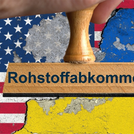 Ein symbolischer Holzstempel mit der Aufschrift "Rohstoffabkommen",vor den Nationalflaggen der Vereinigten Staaten von Amerika und der Ukraine