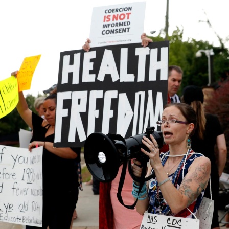 Eine Demonstrantin spricht in ein Megafon. Hinter ihr halten Leute ein Banner mit der Aufschrift "Health Freedom".