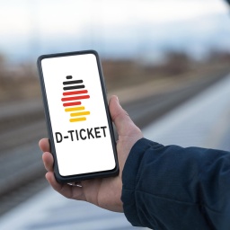 Eine Person steht an einem Bahnsteig und hält in der Hand ein Smartphone in der Hand, auf dem ein Deutschlandticket ist.